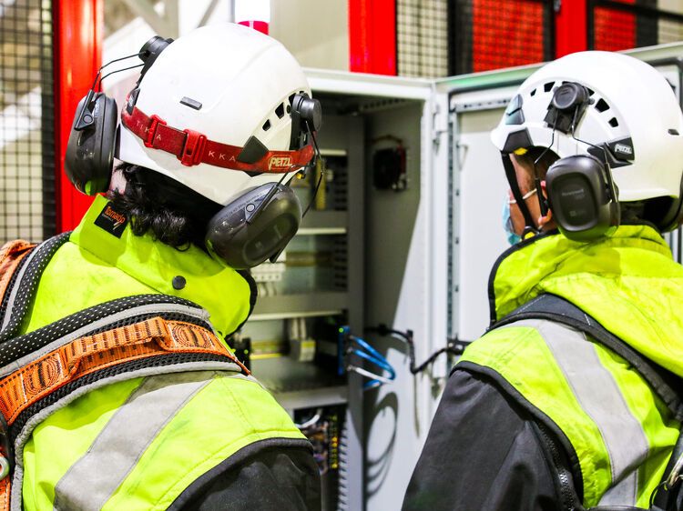 Two technicians in hard hats and harnesses inspecting an open switchboard cabinet