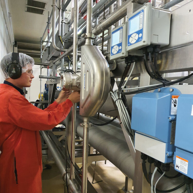Technician in a hairnet and hearing protection adjusting a flow meter on dairy processing pipework